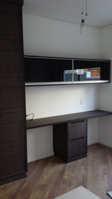 Mesa para escritório com gaveteiro em MDF e armário em MDF com portas basculantes com caixilho de alumínio com vidro preto.